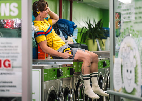 Man sitting on top of laundry machine wearing headphones, a yellow shirt, grey lounge shorts and classic crew socks in black stripe.