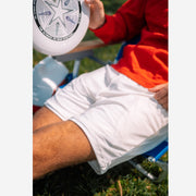 Man sitting on a chair holding a frisbee wearing a red shirt and cream lounge shorts.
