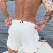 Shirtless man wearing cream lounge shorts standing on boat looking at ocean.