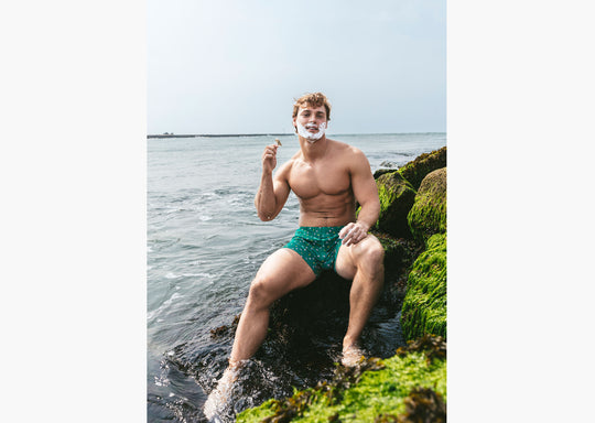 Shirtless man sitting outside by ocean shaving wearing a green Doghouse slim fit boxer (green  with small icons like dog house, golf clubs, card etc)