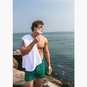Shirtless man standing outside by ocean shaving wearing a green Doghouse slim fit boxer (green  with small icons like dog house, golf clubs, card etc)