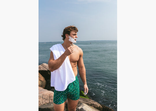 Shirtless man standing outside by ocean shaving wearing a green Doghouse slim fit boxer (green  with small icons like dog house, golf clubs, card etc)