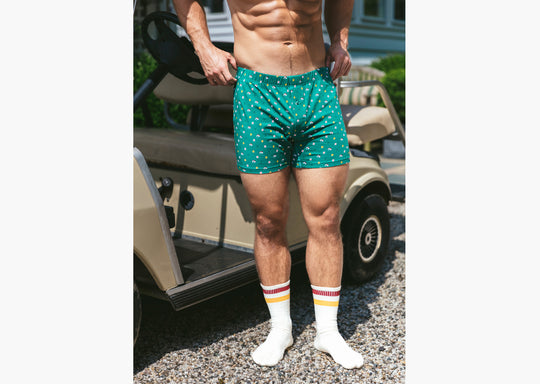 Close up short of shirtless man standing in front of a golf cart wearing a green Doghouse slim fit boxer (green  with small icons like dog house, golf clubs, card etc)