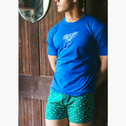 Man wearing a blue shirt and green Doghouse slim fit boxer (green  with small icons like dog house, golf clubs, card etc) standing in front of wooden wall