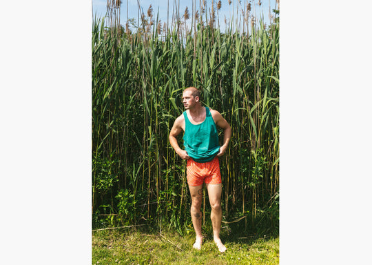 Man standing outside wearing green sleeveless top and orange Doghouse slim fit boxer (orange with small icons like dog house, golf clubs, card etc)
