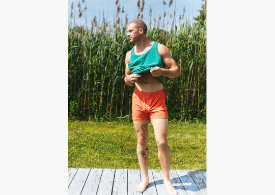 Man standing outside wearing green sleeveless top (lifting it up in photo) and orange Doghouse slim fit boxer (orange with small icons like dog house, golf clubs, card etc)