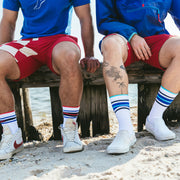 Close up shot of two men sitting on wood pier at the beach wearing fieldhouse striped crew socks and red lounge shorts.