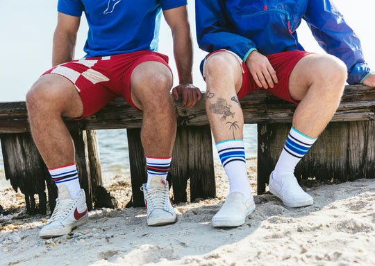 Close up shot of two men sitting on wood pier at the beach wearing fieldhouse striped crew socks and red lounge shorts.