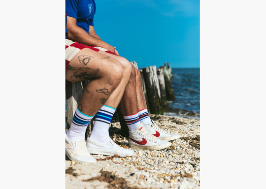 Close up side shot of two men sitting on wood pier at the beach wearing fieldhouse striped crew socks and red lounge shorts.