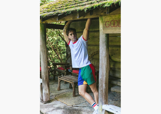 Man standing in front of wood cabin wearing white t shirt and blue/green/yellow/red mesh shorts.