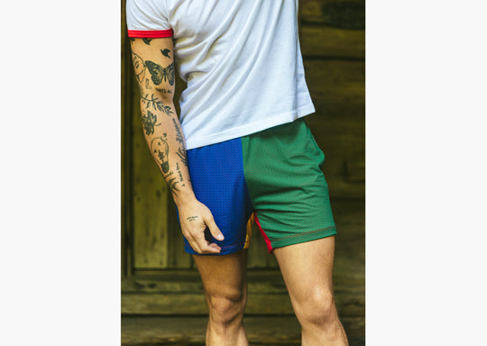 Close up of man standing in front of cabin wearing white t-shirt and blue/gree/yellow/red colored mesh lounge shorts.
