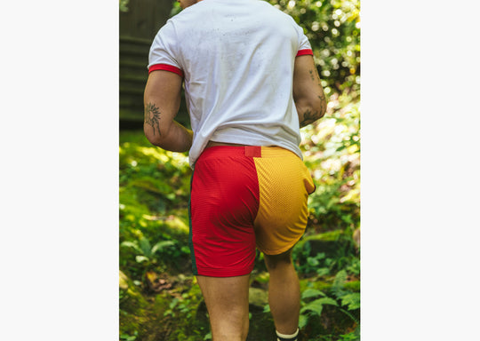 Close up back shot of man walking through forest wearing white t shirt and red/yellow colored mesh shorts.