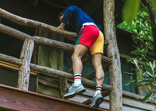 Man climbing onto railing of wood cabin wearing navy hoodie and green/red/yellow colored mesh shorts.