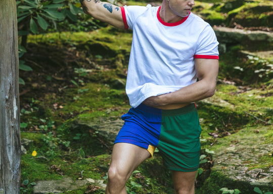 Man standing in forest wearing white t-shirt and blue/green/yellow colored mesh shorts.