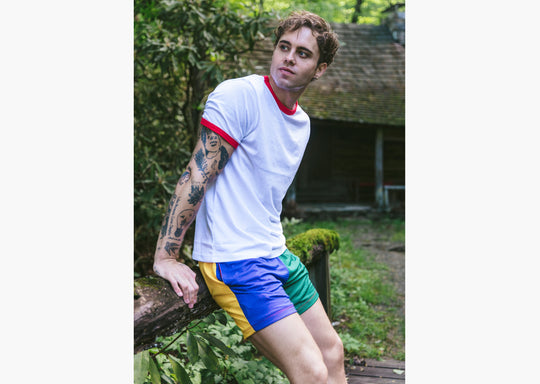 Man leaning against wooden railing wearing white t shirt and yellow/blue/green mesh lounge shorts. 