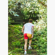Back shot of man walking up stone steps in forest wearing white t shirt and red/yellow colored lounge shorts.