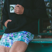 Man holding coffee sitting at table wearing black hoodie and green madras slim fit boxer