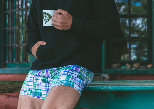 Man holding coffee sitting at table wearing black hoodie and green madras slim fit boxer