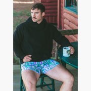Man holding coffee sitting at table wearing black hoodie and green madras slim fit boxer