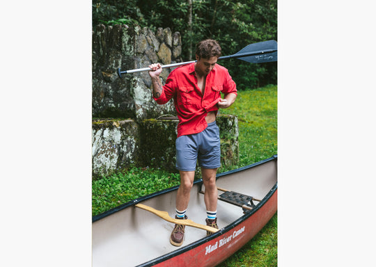 Man standing inside of canoe with paddle over shoulder weariing red shirt and grey lounge shorts.