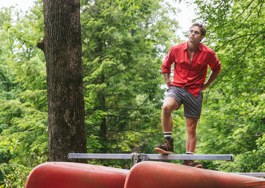 Man in forest standing on stack of canoes wearing a red shirt and grey lounge shorts.