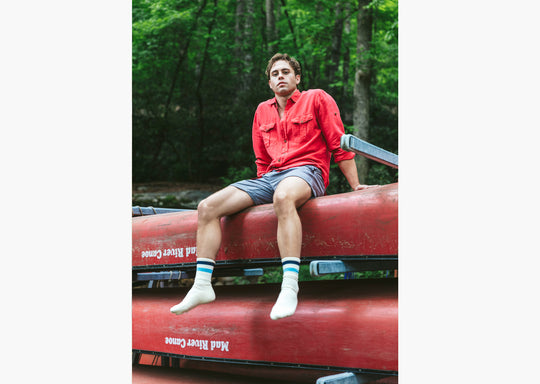 Man in forest sitting on top of pile of canoes wearing red shirt and grey lounge shorts.