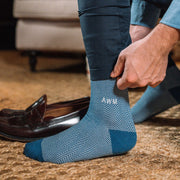 Close up shot of man getting dressed wearing blue herringbone dress socks with AWM initials monogrammed on one sock.