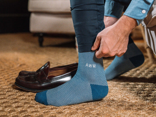 Close up shot of man getting dressed wearing blue herringbone dress socks with AWM initials monogrammed on one sock.