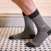 Man standing inside bathroom wearing black herringbone dress socks with AWM initials monogrammed on one sock. 