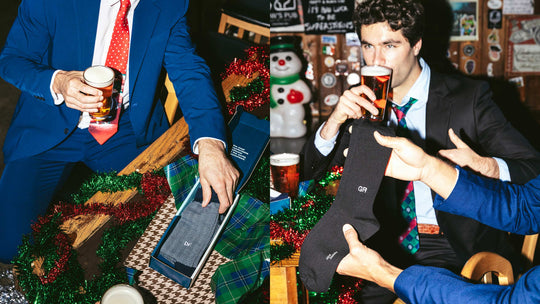 Men in suits at a holiday-decorated bar with boxed patterned socks and black GR monogram socks held up for display.