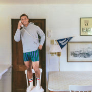 A man in a gray hoodie and green striped boxer briefs stands on a white chair in a bright room, holding a vintage white telephone to his ear.