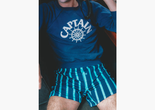 Close up of man sitting on boat holding radio wearing navy sweatshirt and navy stripe slim fit boxers.
