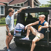 Two men in lounge shorts and t shirts hanging out leaning on a Jeep in a driveway.