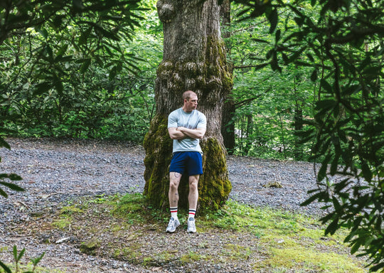 Man standing in forest with arms crossed wearing grey tshirt and navy blue lounge short.