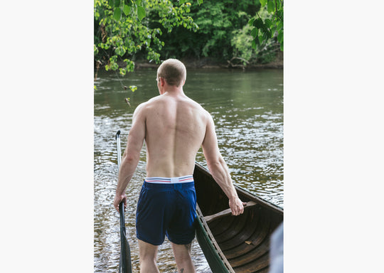 Back shot of a shirtless man pulling a canoe in water holding a paddle and wearing navy blue lounge shorts.