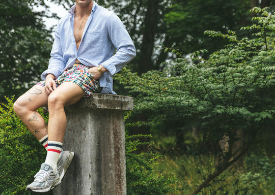 Man sitting in forest on cement pillar wearing Madras print lounge short, blue opened dress shirt, and white/red/blue crew socks.