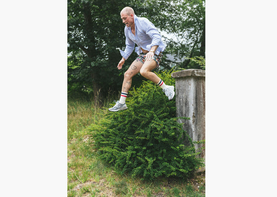 Man in forest jumping off cement pillar wearing blue dress shirt, madras printed lounge short, and red/white/blue crew socks.