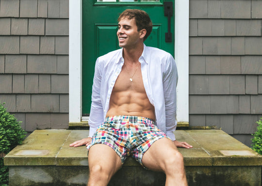 Man sitting on house porch wearing opened dress shirt and Madras printed lounge shorts.