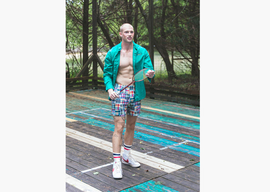 Full body shot of man wearing green dress shirt and Madras print lounge shorts standing on wooden court holding a racket.