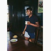 Man wearing blue shirt and red/white/blue Regatta slim fit boxer standing at a table with waffles and holding a coffee..