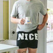 Man in kitchen holding coffee mug and wearing a pair of black slim fit boxers with the words "NICE" in big bold white font.