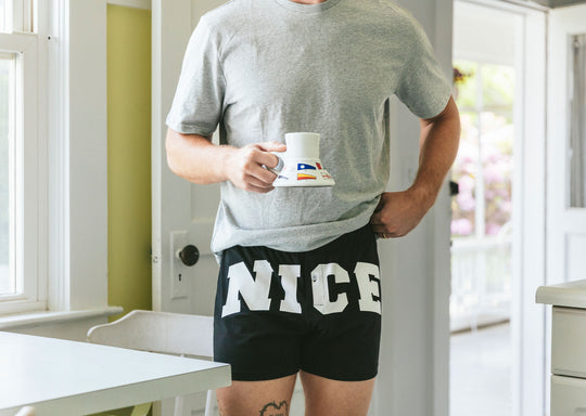 Man in kitchen holding coffee mug and wearing a pair of black slim fit boxers with the words "NICE" in big bold white font.