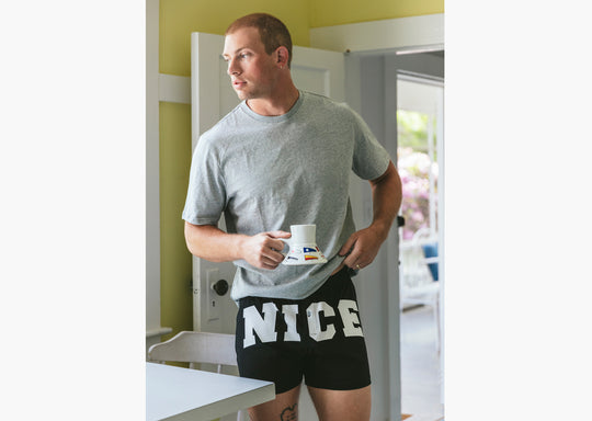 Man in kitchen holding coffee mug and wearing a pair of black slim fit boxers with the words "NICE" in big bold white font.