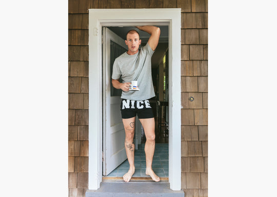 Man in doorway holding coffee mug and wearing a pair of black slim fit boxers with the words "NICE" in big bold white font.