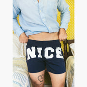 Close up of man in bedroom with yellow walls and he is wearing blue slim fit boxers with words "NICE" in big bold white font. 