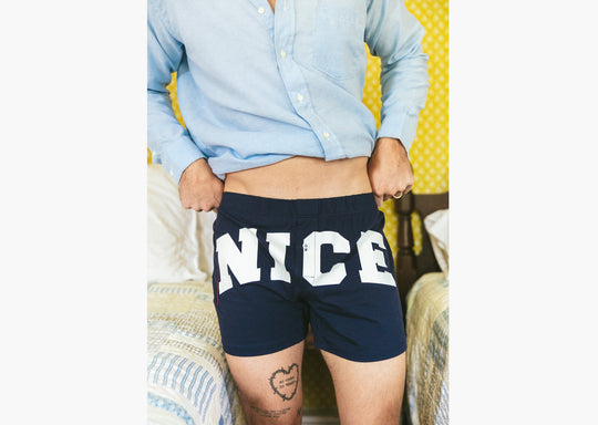 Close up of man in bedroom with yellow walls and he is wearing blue slim fit boxers with words "NICE" in big bold white font. 