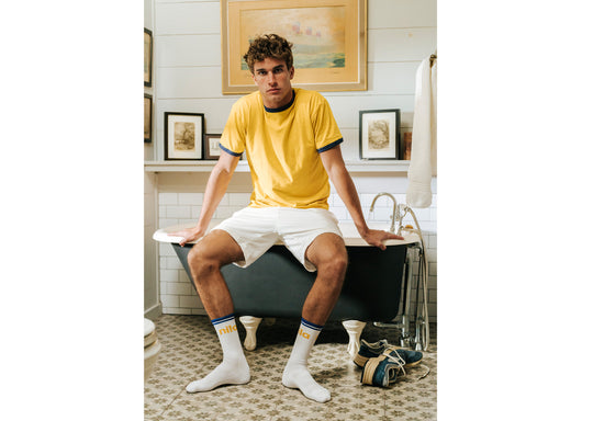 Man sitting on edge of bath wearing yellow shirt, white shorts and white/yellow/blue crew socks with letters NILA on them