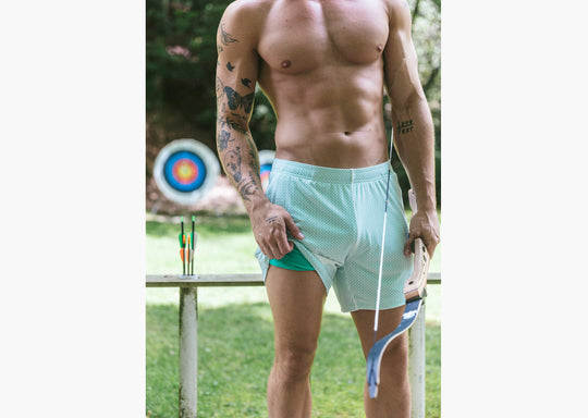 Shirtless man standing in archery range holding a bow and wearing white/green mesh lounge shorts. The man is lifting the shorts to show the green interior.