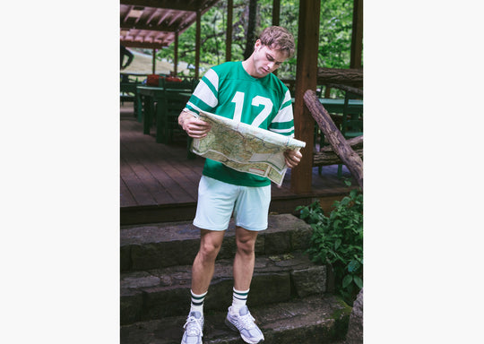 Man standing on stone steps wearing green/white jersey with number 12 holding a map and also wearing white lounge shorts.