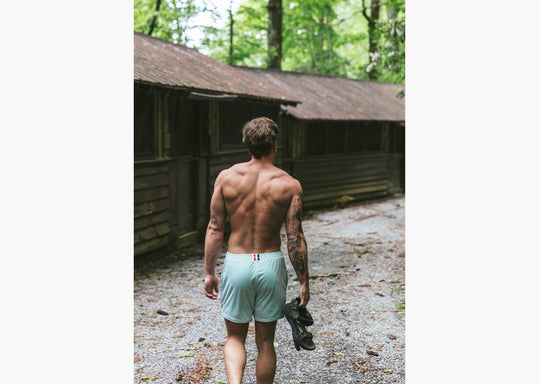 Back short of man walking next to cabins holding sandals and wearing white mesh shorts.